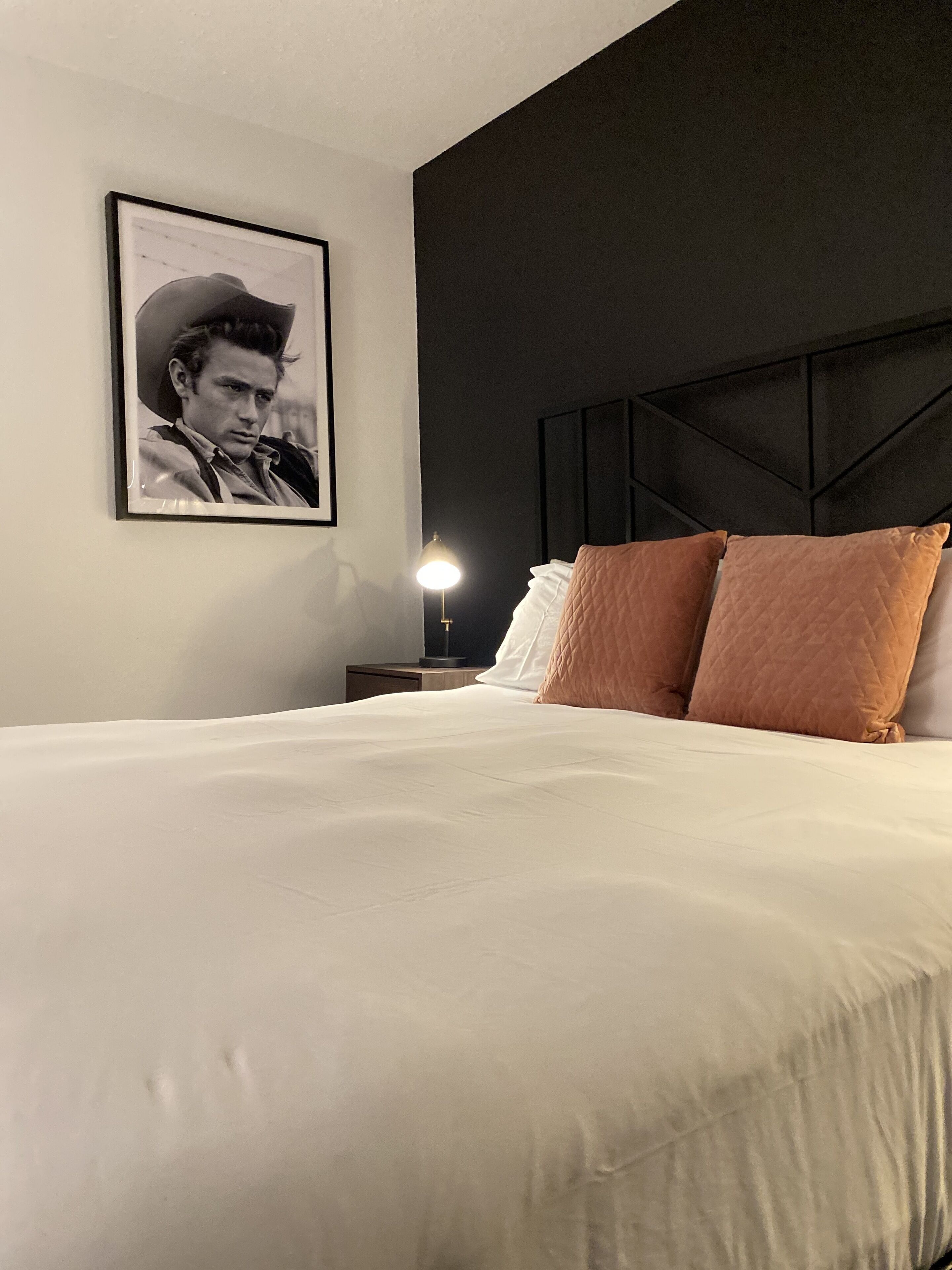 Cama bem arrumada com edredom branco e dois travesseiros, quadro na parede e abajur na mesinha de cabeceira.