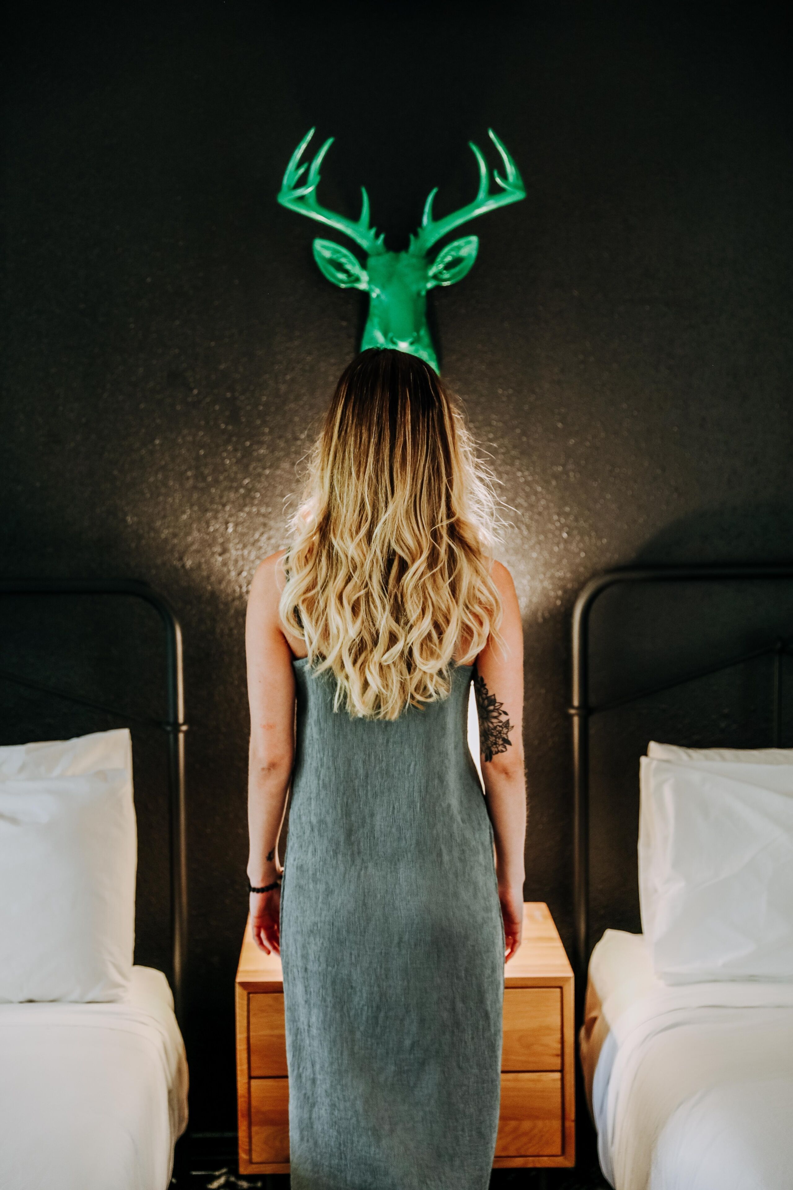 A person in a grey dress standing in a room with two beds, each with white bedding and wooden headboards.
