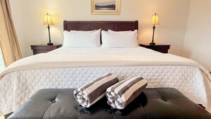 Una cama bien hecha con ropa blanca, dos lámparas de noche, un cuadro enmarcado en la pared y un banco de cuero negro con toallas.