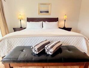 A neatly made bed with white bedding, two bedside lamps, a framed picture on the wall, and a black leather bench with towels.