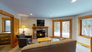 A cozy living room with a fireplace, a television, and a large window offering a view of trees. - Hibernation House at Lutsen, Hibernation House at Lutsen (Lutsen)