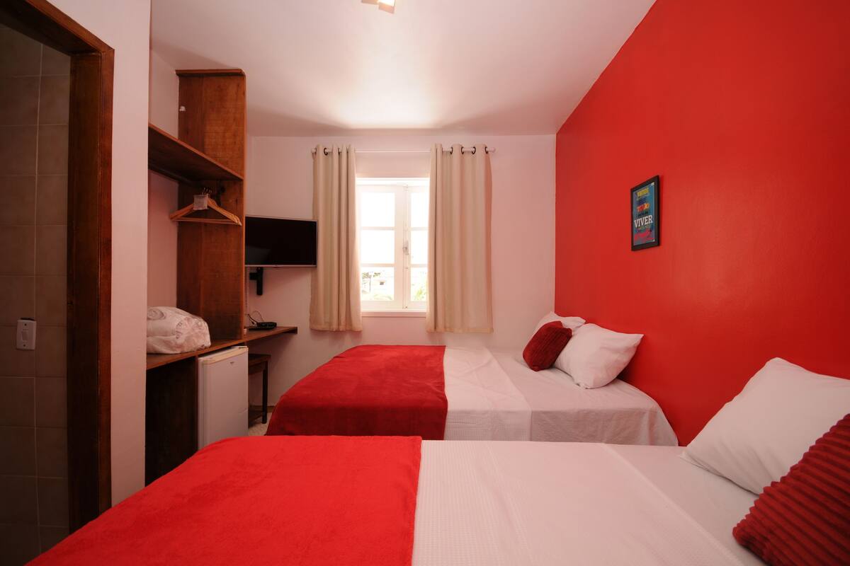 A room with two beds, a wooden wardrobe, a small desk, a television, and a window with curtains.