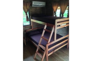 A bunk bed with a wooden frame and a mattress in a cabin-style room with a window and curtains. - Rear Cabin (2 Adults) (Uvita)