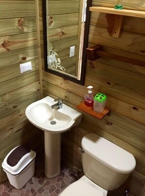 A small bathroom with wooden walls, a white sink, a toilet, and a mirror. - Rear Cabin (2 Adults) (Uvita)