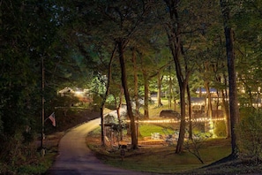 A nighttime view of a forested area with a paved path leading to a building with string lights.