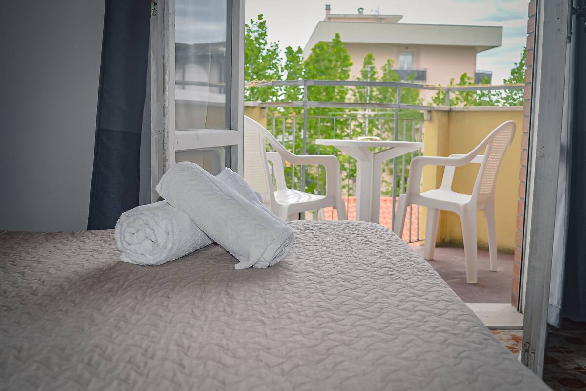 Quarto com cama, toalha enrolada e varanda com cadeiras e mesa.