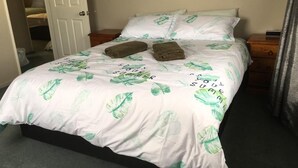 A neatly made bed with a tropical leaf pattern bedspread, two folded towels, and a wooden nightstand with a small lamp. - Irishman Drive Retreat, free wifi. (Twizel)
