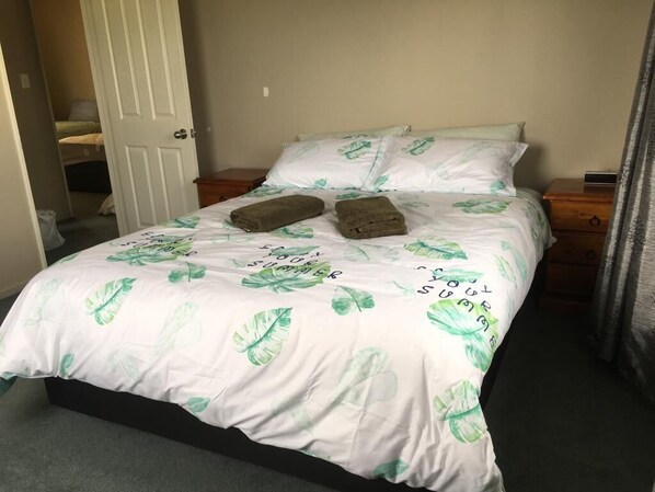 A neatly made bed with a tropical leaf pattern bedspread, two folded towels, and a wooden nightstand with a small lamp.