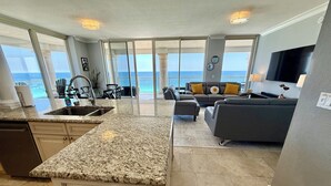 Eine moderne Küche mit einer Granitabdeckung und einem Essbereich mit grauem Sofa und gelben Kissen. Ein großes Fenster bietet Blick auf den Ozean.
