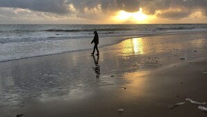 Ein Strand mit einer Person, die am feuchten Sand entlanggeht, Wellen im Hintergrund und einem bewölkten Himmel, während die Sonne untergeht.