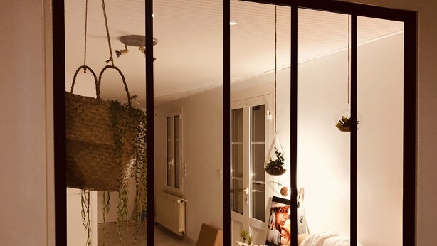 Ein Schlafzimmer mit großem Fenster, einer hängenden Pflanze und einem Bett mit Kissen und Decke.
