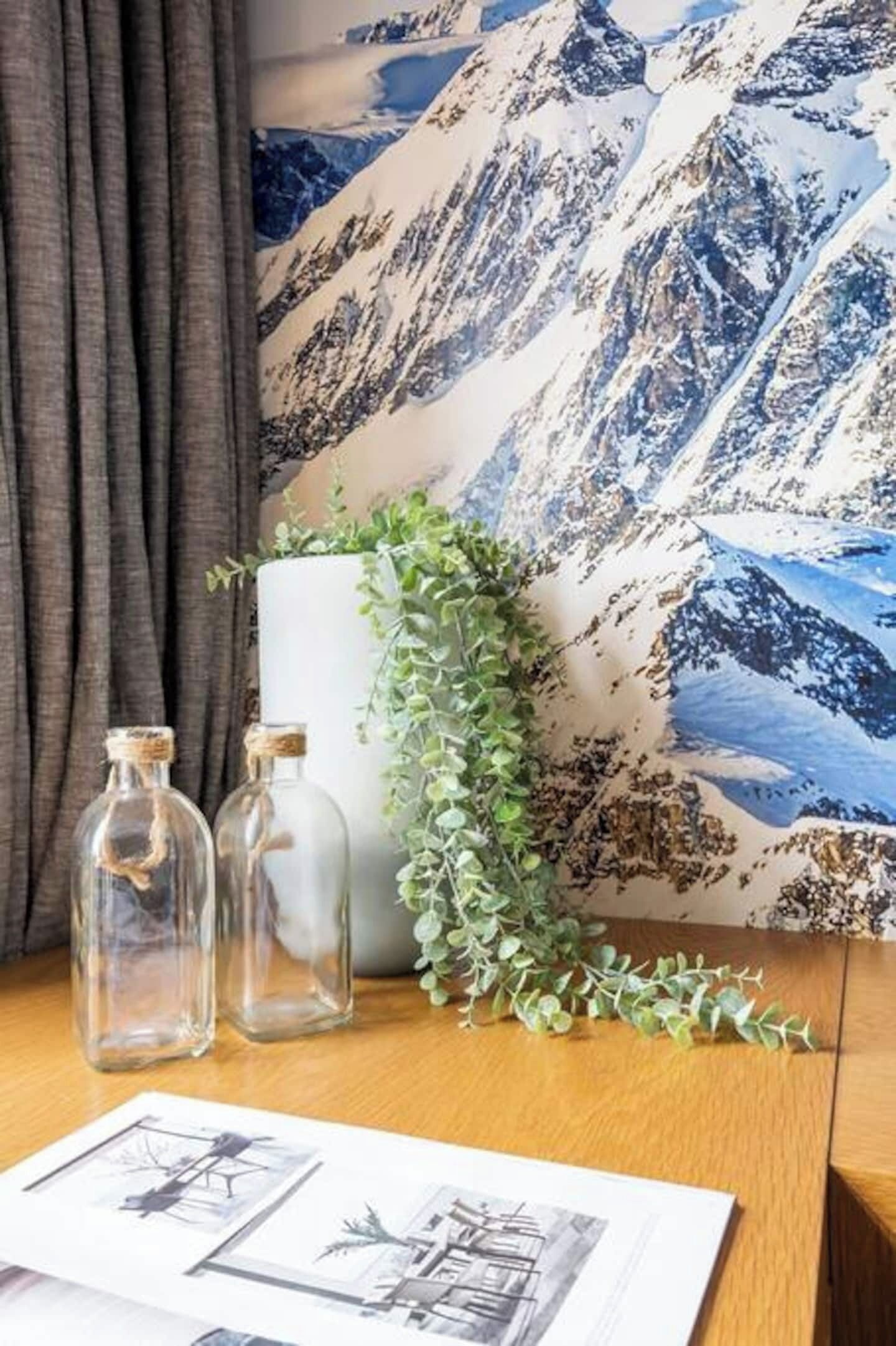 Um quarto com parede decorada com tema de montanha, um vaso branco com folhagem verde e duas garrafas de vidro transparente com rolhas de cortiça.