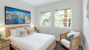 Ein Schlafzimmer mit einem großen Bett, zwei Nachttischen, einem Sessel und zwei Fenstern.