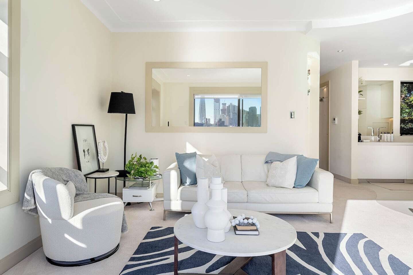 A modern living room with a white sofa, a round coffee table, and a city view through a large mirror.
