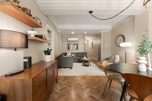 A modern living room with a herringbone floor, a grey sofa, a dining table with chairs, and a kitchen area with wooden shelves.