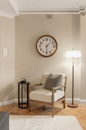 A room with a wooden chair, a round wall clock, a floor lamp, and a small table with a decorative item.