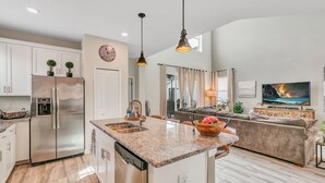 A modern kitchen with a central island, stainless steel appliances, and a dining area with a sofa and TV. - 5BD/5BA Gorgeous Home w/ pool close to Disney (Kissimmee)