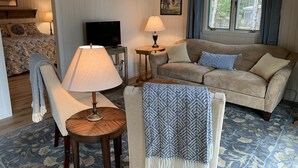 Una sala de estar acogedora con un sofá, un sillón y una mesita con una lámpara. Un televisor está sobre un mueble bajo, y hay dos cuadros en la pared.