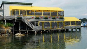 Un edificio frente al agua con techo amarillo y varios niveles, que cuenta con mesas al aire libre y jardineras con flores.