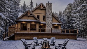 Un chalet doté d’une cheminée en pierre, d’une terrasse en bois et de chaises recouvertes de neige.