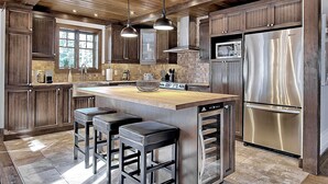Une cuisine avec des meubles en bois, un réfrigérateur en acier inoxydable, un grand îlot central avec des tabourets, des luminaires suspendus et une fenêtre donnant sur les arbres.