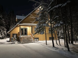 A two-story wooden cabin with a snow-covered porch and steps leading to the entrance.