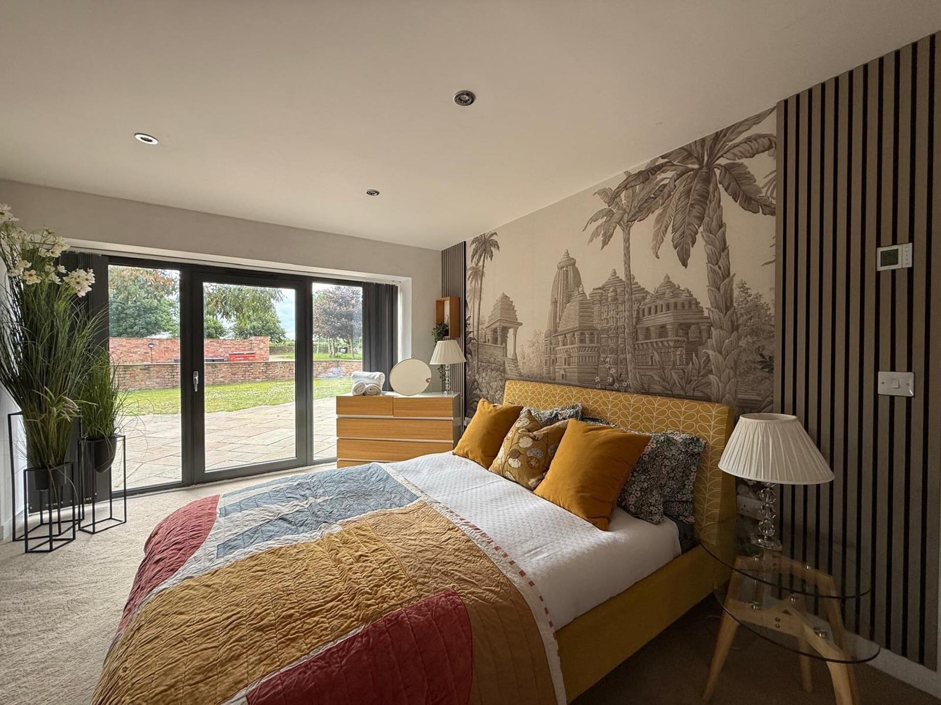 A bedroom with a large bed, a view through the glass door, a wall mural, and a potted plant.