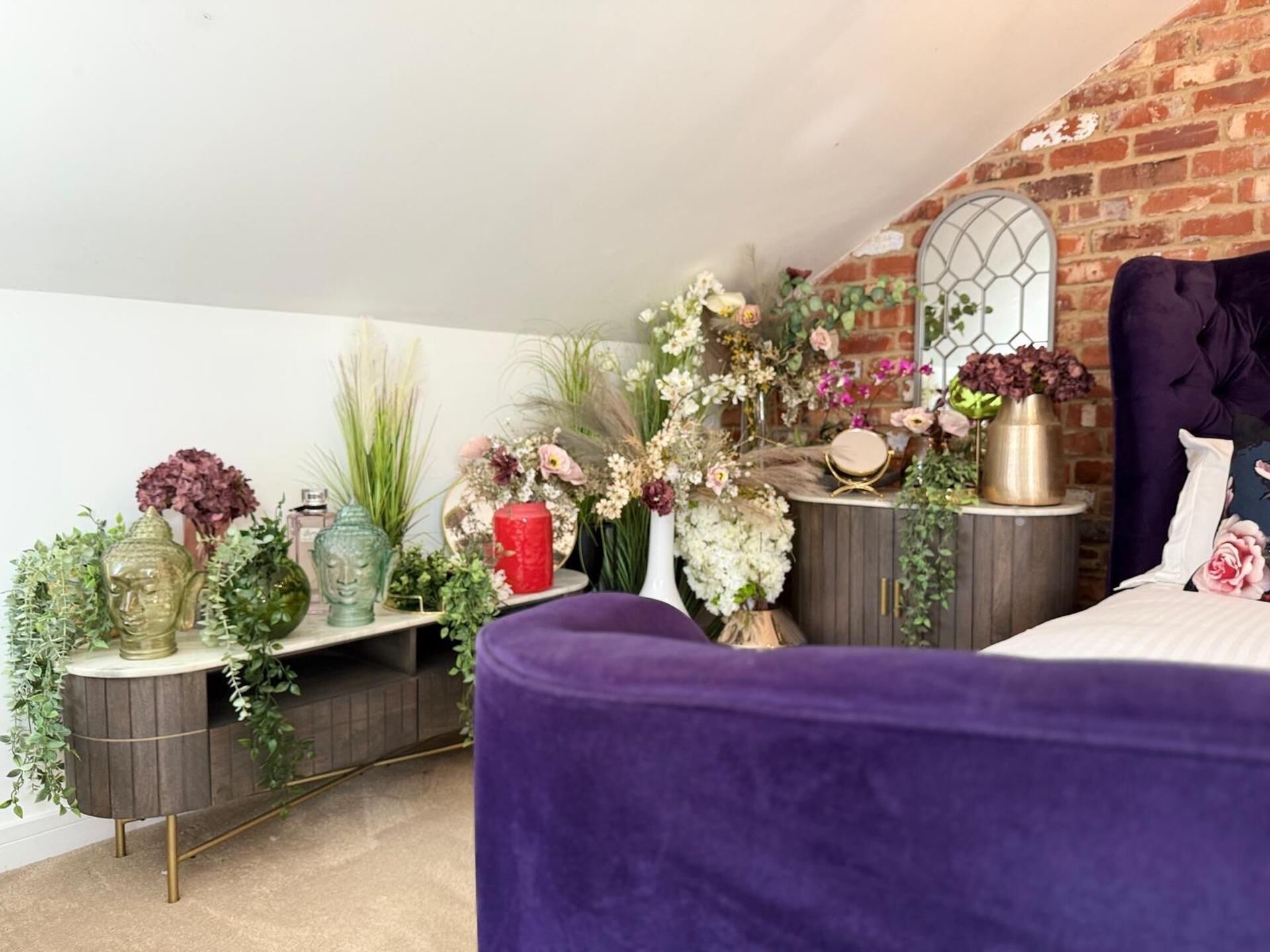 A bedroom with a purple bed, a brick wall, and various decorative items including plants and vases.
