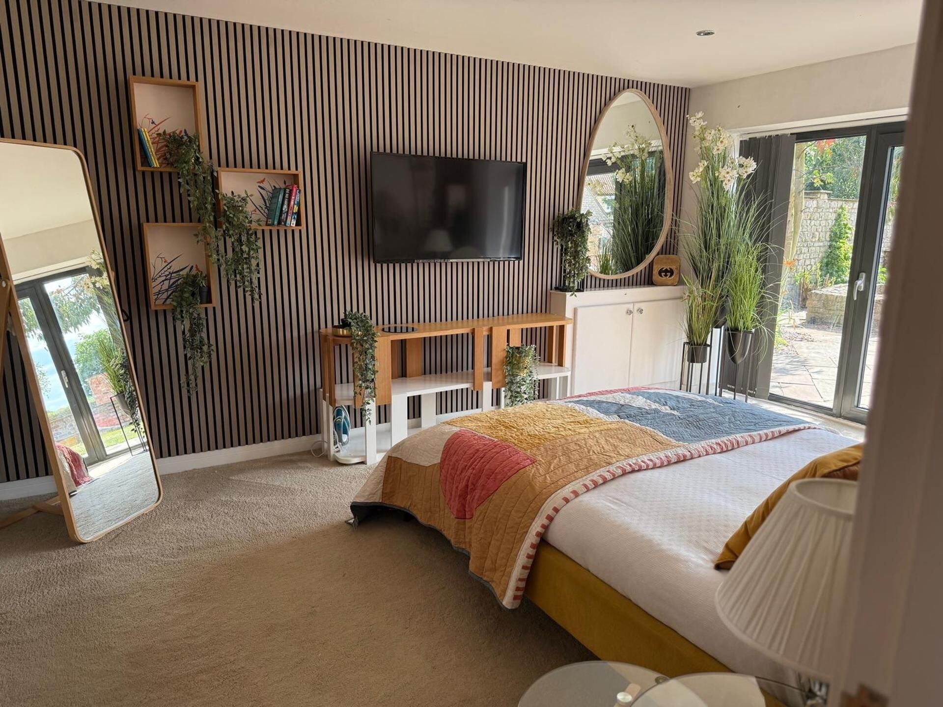 A modern bedroom with a large bed, a TV, a mirror, and a window with plants.