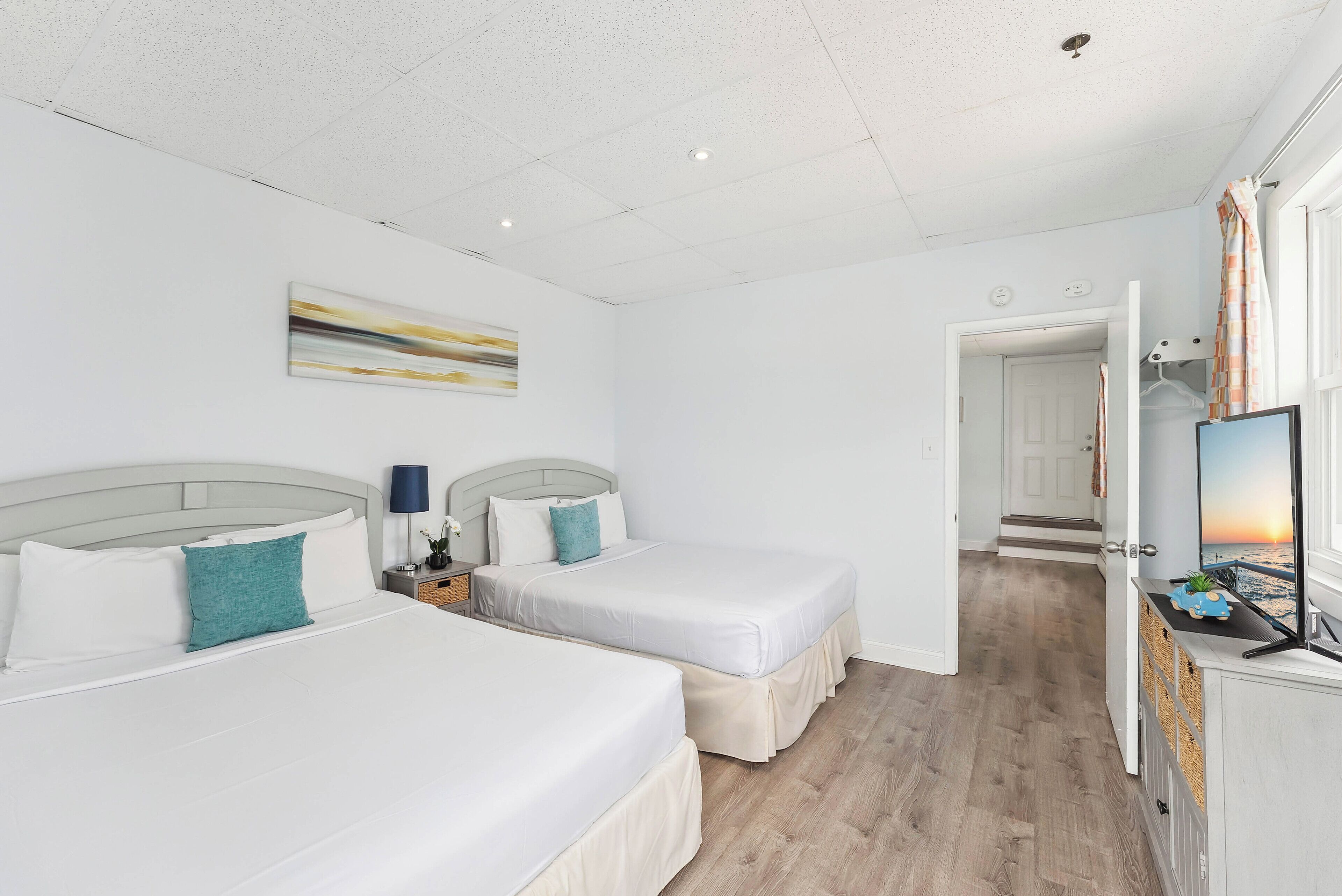 A hotel room with two beds, a nightstand, a lamp, a painting, and a view of the sunset through a window.