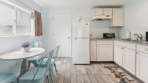 A compact kitchen with white cabinets, a refrigerator, a dining table with chairs, and a window with curtains. - Daytona Inn and Suites - Efficiency Queen (Wildwood)