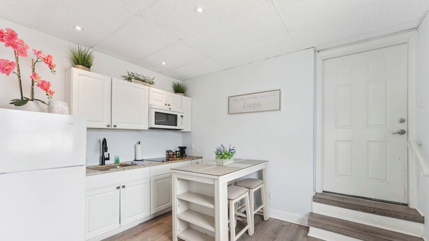 A kitchen with white cabinets, a microwave, a sink, and a small table with two stools. There is a door with a sign above it. - Daytona Inn and Suites - Efficiency Queen (Wildwood)