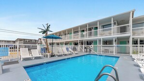 A pool area with lounge chairs, a blue umbrella, and a multi-story building with balconies. - Daytona Inn and Suites - Efficiency Queen (Wildwood)
