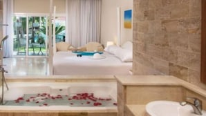A hotel room with a large bed, a bathtub with rose petals, a shower, and a view of a pool and palm trees. - Junior Suites – The Tropical (Puerto Vallarta)