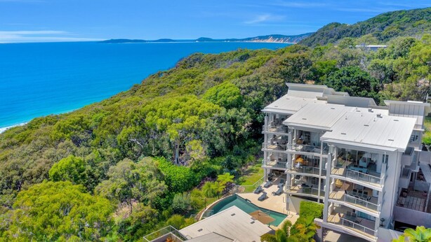 A modern multi-story hotel with a pool, surrounded by lush greenery and overlooking a coastal area.