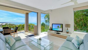 A modern living room with a large TV, comfortable seating, and a balcony with a scenic ocean view.