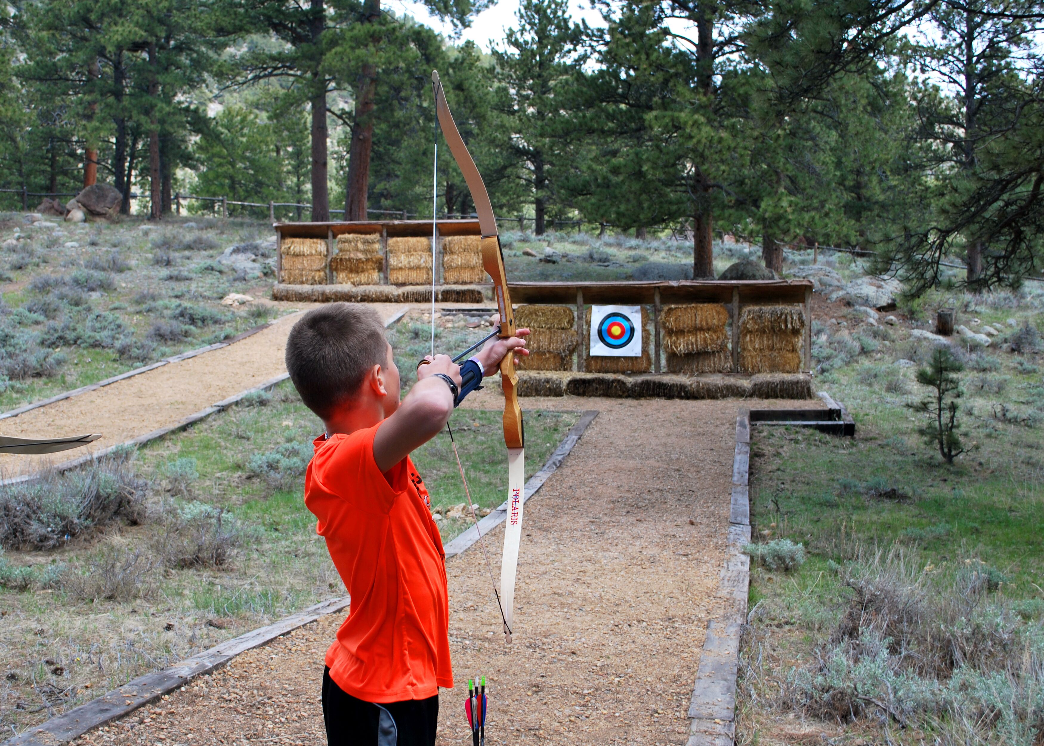 A person aiming a bow at a target in an outdoor setting with trees and hay bales.