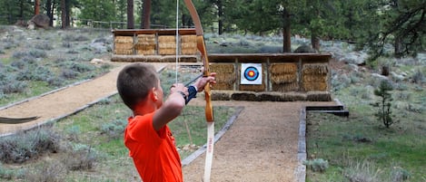 A person aiming a bow at a target in an outdoor setting with trees and hay bales.
