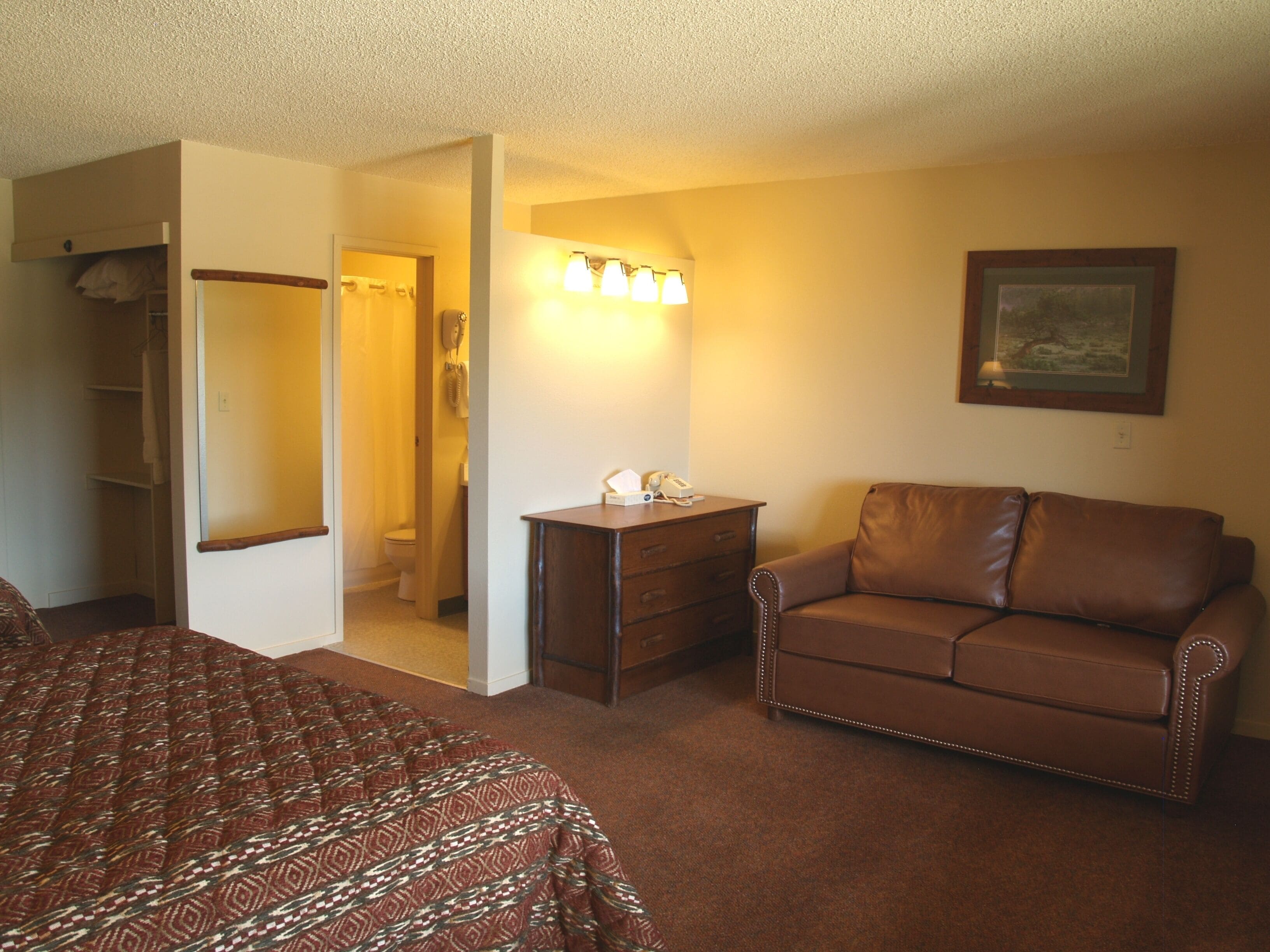 A hotel room with a bed, a brown leather sofa, a dresser, and a bathroom visible through an open door.
