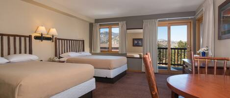 A hotel room with two beds, a dining table, and a balcony with a view of the mountains.
