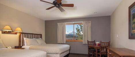 A hotel room with two beds, a dining table with chairs, a ceiling fan, and a window with curtains.