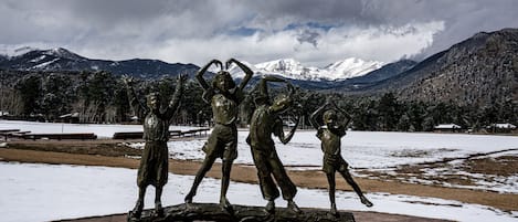 A sculpture of three figures on a log in a park with snow and mountains in the background.