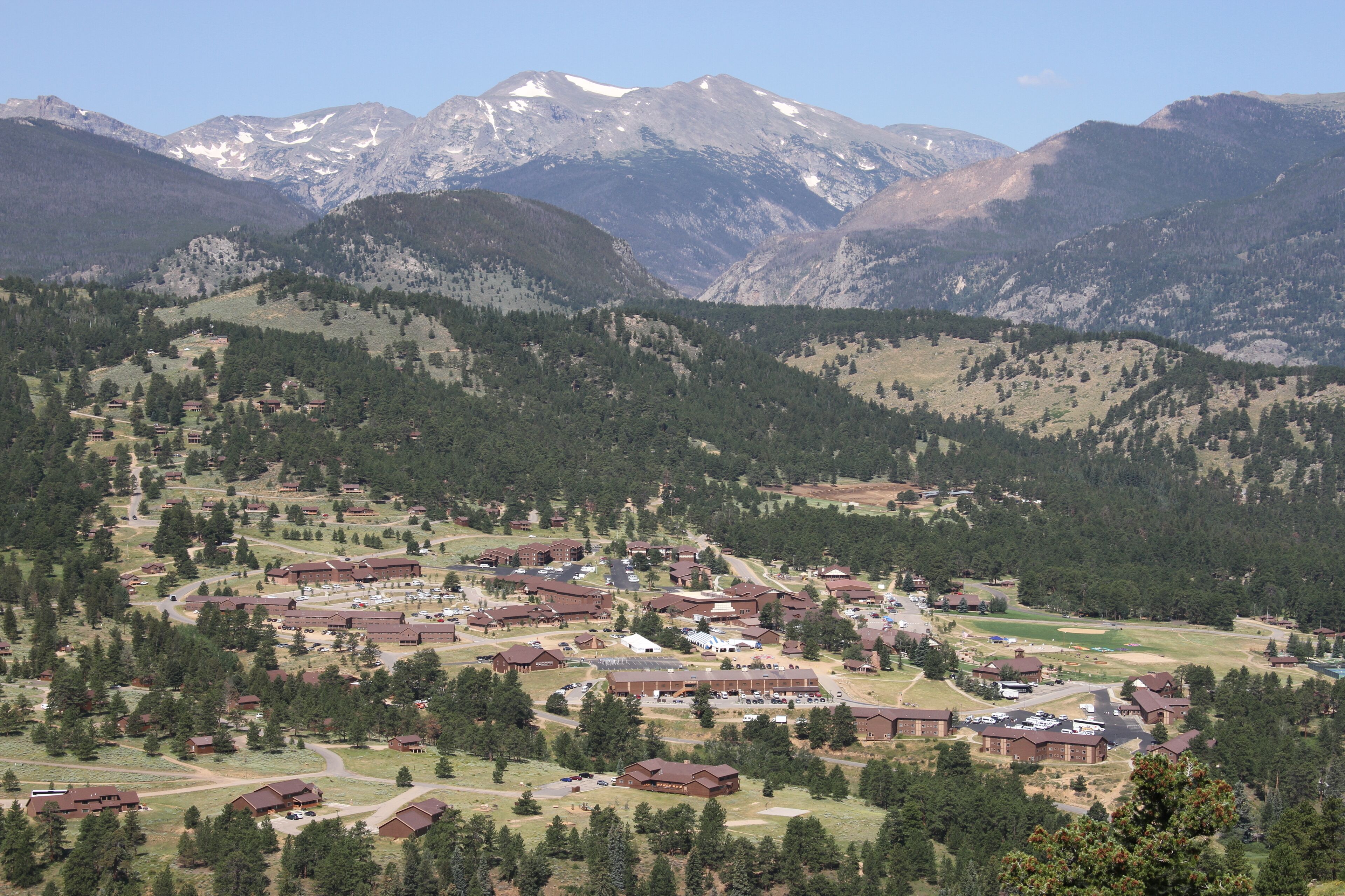 Uma região montanhosa com um conjunto de edifícios, provavelmente um resort ou pousada, rodeada por densas florestas e picos nevados ao fundo.