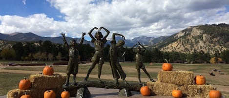 Una escultura de niños sobre un tronco, con fardos de heno y calabazas en un parque con montañas al fondo.