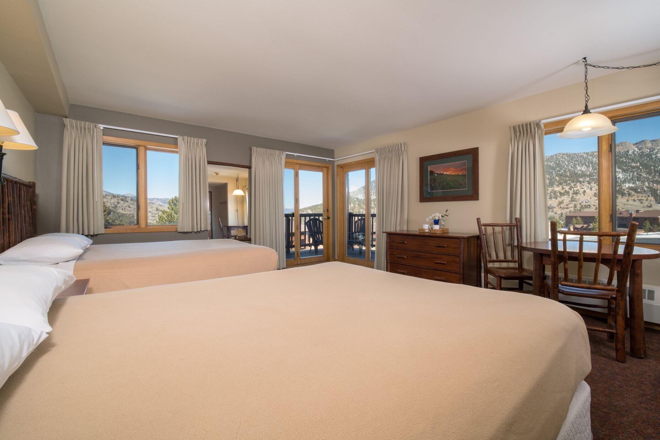 A hotel room with two beds, a desk, chairs, and large windows offering a mountain view.