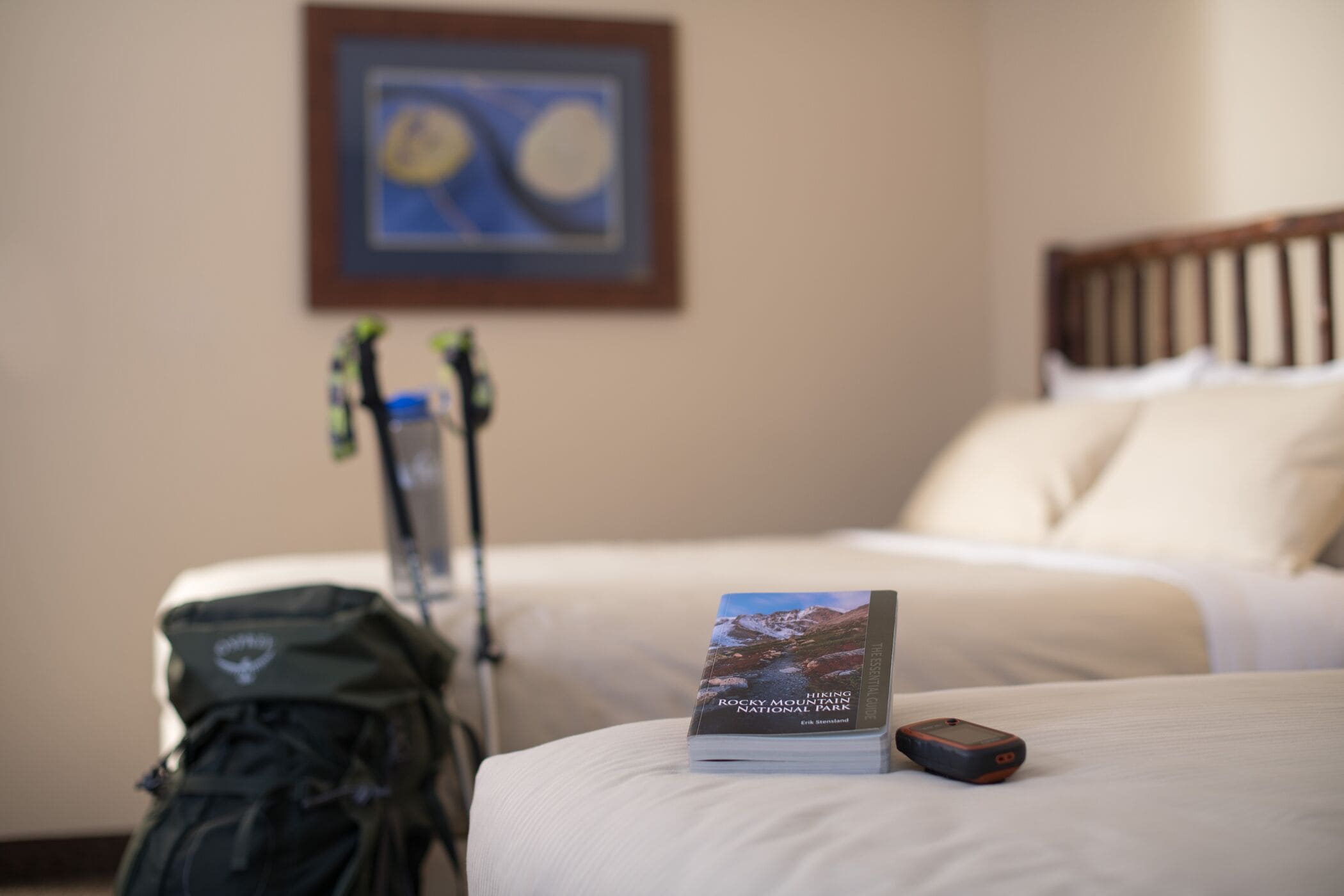 Una habitación de hotel con una cama, un libro titulado "Hiking the Rocky Mountains National Park", una mochila y un par de esquíes apoyados contra la pared.