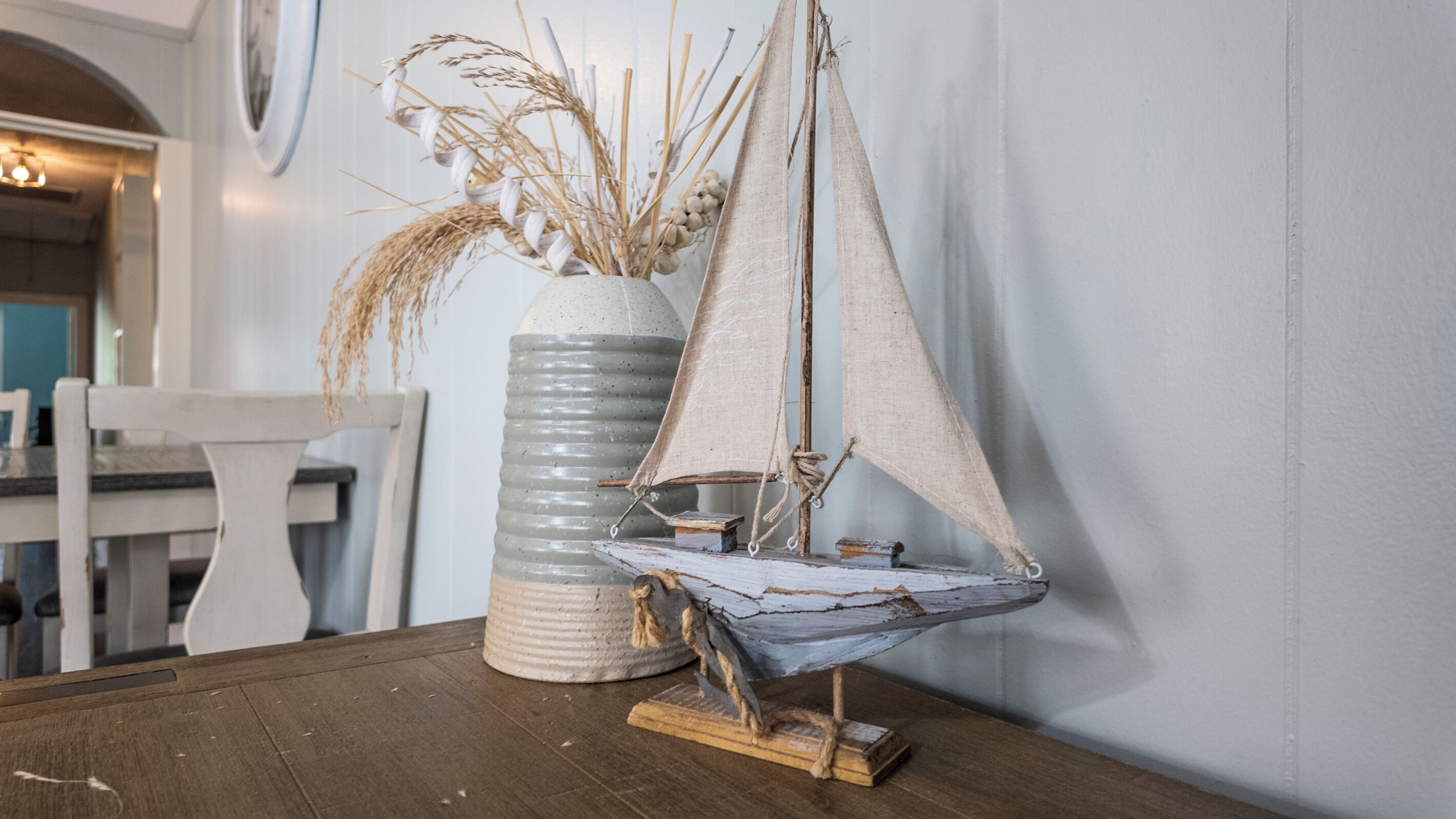A rustic interior with a wooden table, a white vase with dried plants, and a sailboat model.