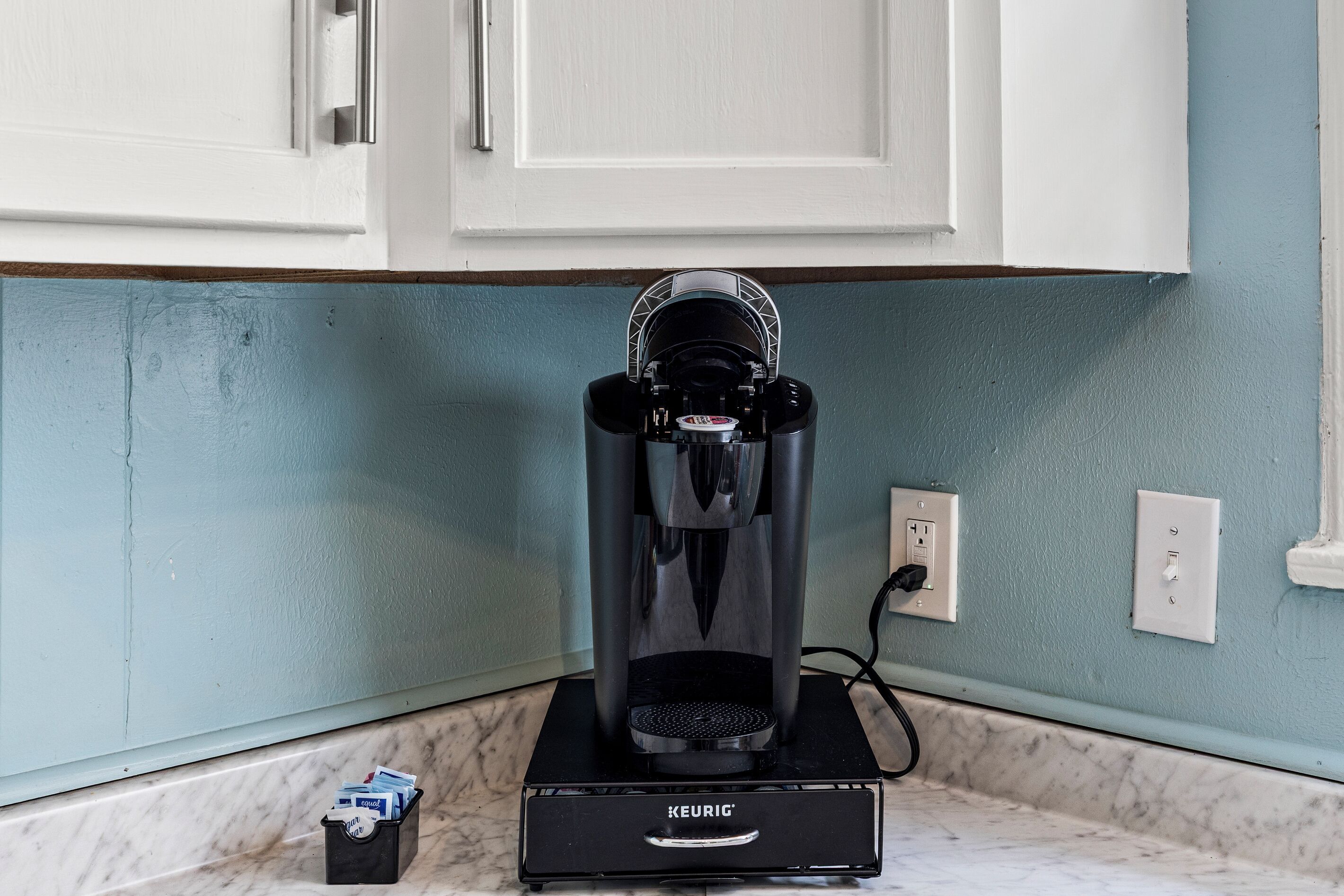 A Keurig coffee machine on a countertop with a white cabinet above and a light blue wall in the background.