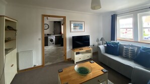 A compact living space with a kitchenette, a TV, a sofa, and a view of the outside. - Little Acorn - 2-bed Anstruther Apartment (Anstruther)