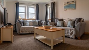 A cozy living room with a sofa, a coffee table, and a television on a stand. - Little Acorn - 2-bed Anstruther Apartment (Anstruther)